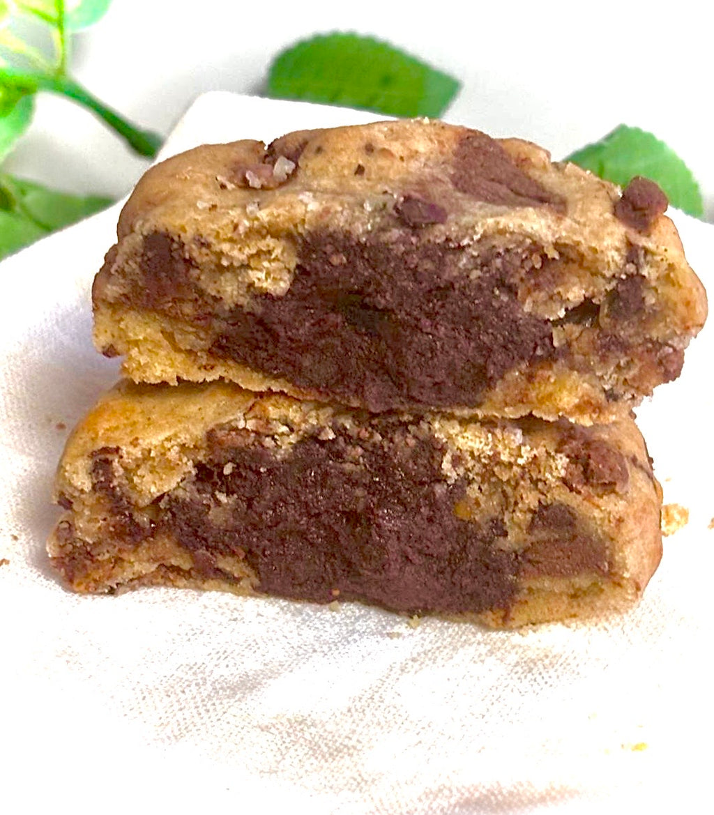 Two chocolate chip cookie stacked on a white cloth with green leaves in the background