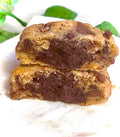 Two chocolate chip cookie stacked on a white cloth with green leaves in the background