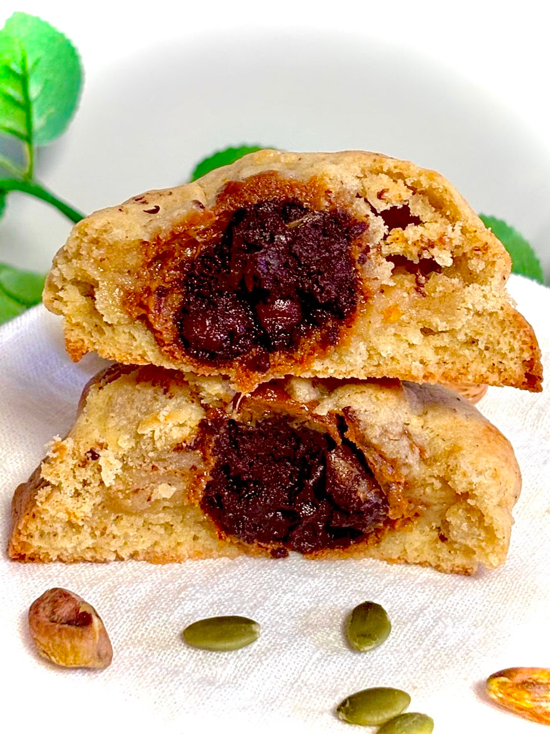 Two halves of a cookie with a chocolate filling on a white surface with green leaves in the background.